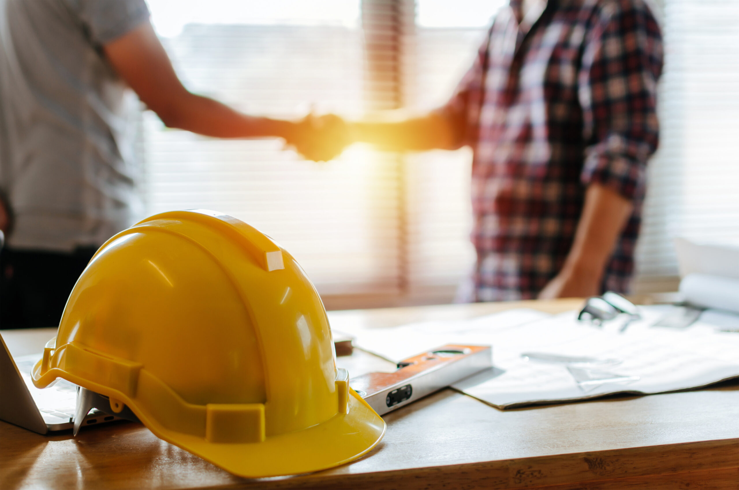photo of yellow safety helmet on workplace desk with construction worker team hands shaking greeting start up plan new project contract in office center at construction site, partnership and contractor concept